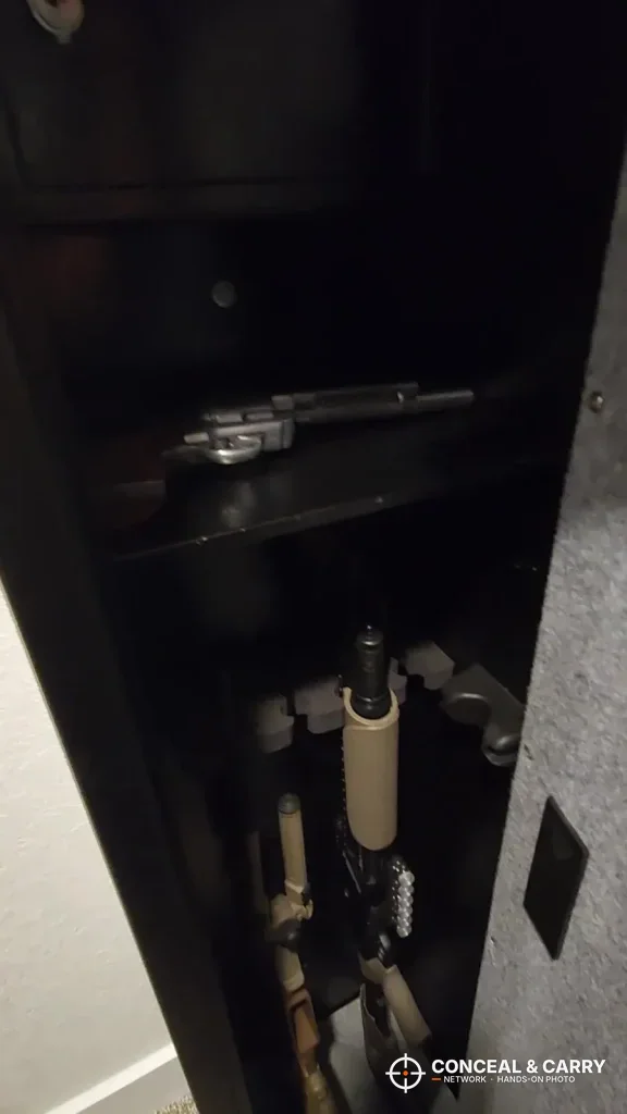 Dim view inside the BBRKIN rifle safe with a pistol on the shelf and long guns below