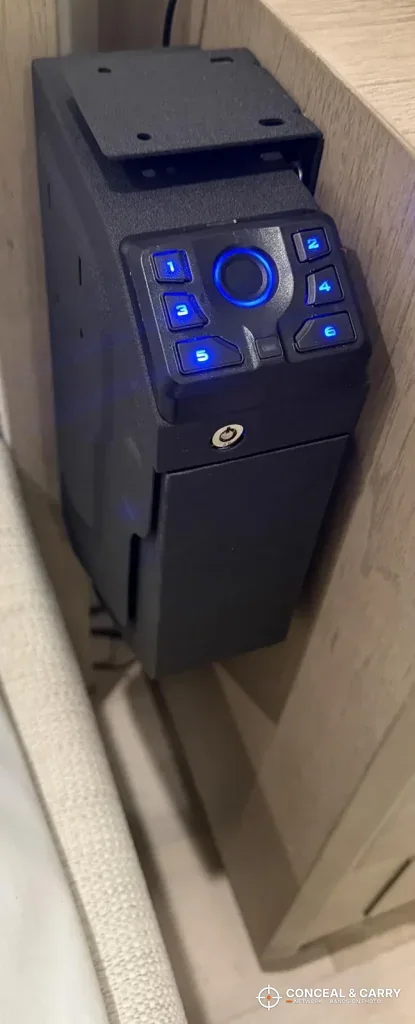 Black VERPROOF gun safe with blue backlit buttons, mounted beside a cabinet